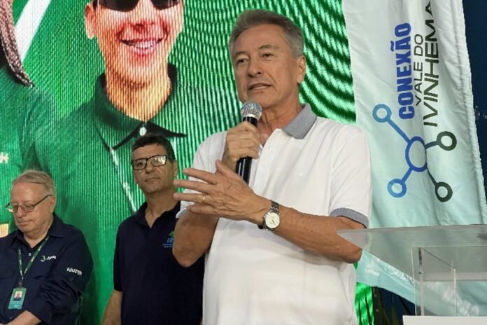 Man in a white polo speaks into a handheld microphone on stage, with two men in the background and a large digital screen behind him.