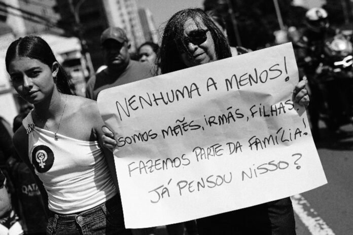 Protester holding a handwritten Portuguese sign at a street rally: 'Nenhuma a menos! Somos mães, irmãs, filhas... Já pensou nisso?'.