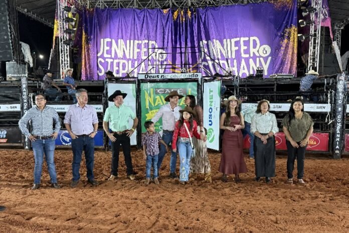 Group of men, women, and two children standing in a dusty arena in front of a large purple stage backdrop.