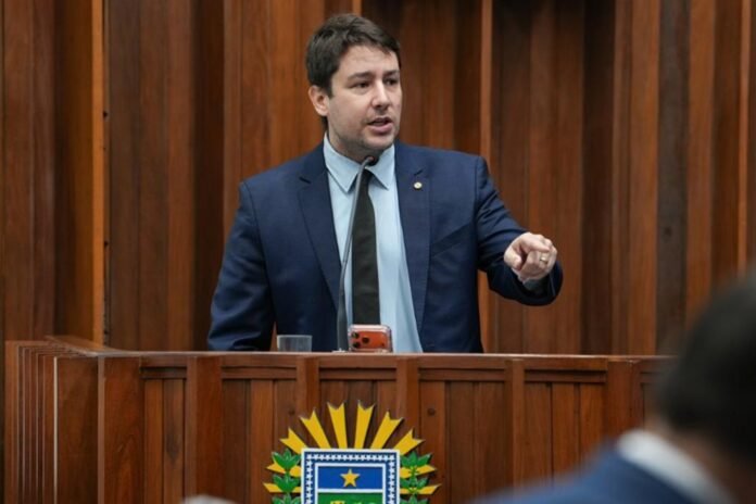 Man in a navy suit speaking at a wooden podium with a microphone in a formal chamber.