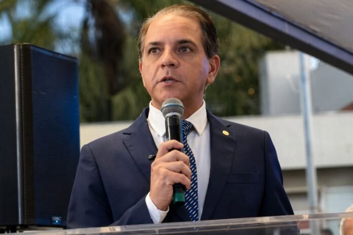Man in a navy suit speaks into a handheld microphone at an outdoor podium.