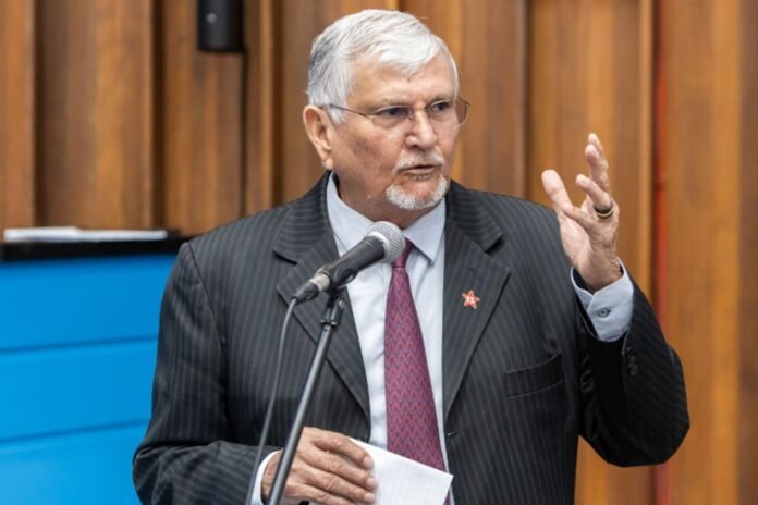 Older man in a pinstripe suit and red tie speaks into a microphone, gesturing with his right hand while holding papers at a podium.