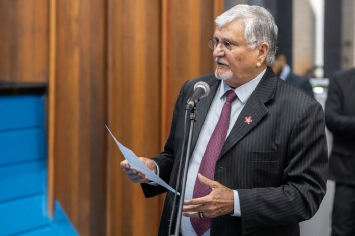 Older man in a pinstriped suit and red tie speaks into a microphone while holding a sheet of paper at a formal event.
