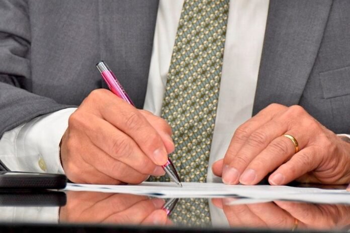 Close-up of a person in a suit signing a document with a pink pen, wearing a wedding ring on the left hand.