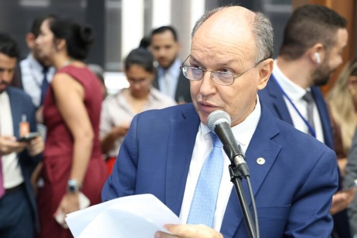 Man in a blue suit and light blue tie speaks into a microphone while holding papers at a formal event.