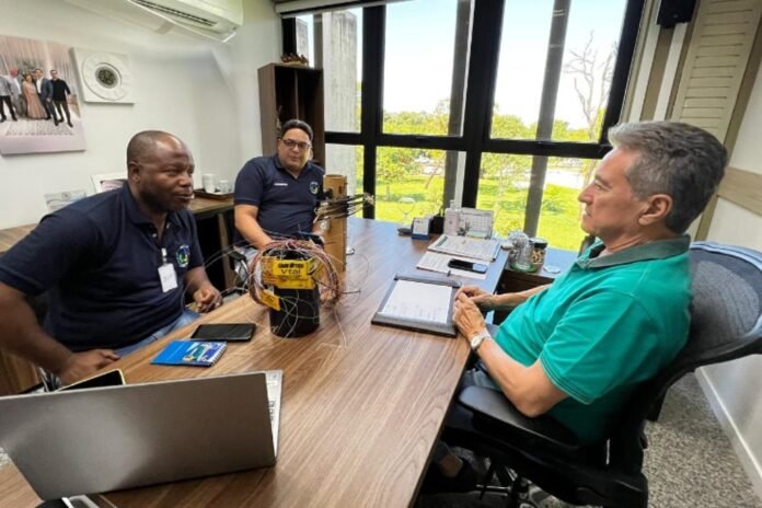 Three men in a meeting around a wooden desk in an office, with a wire harness prototype on the table and laptops open.