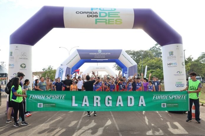 Group of runners at the starting line behind a bright green a 'LARGADA' banner, with purple inflatable arches overhead during a race event.