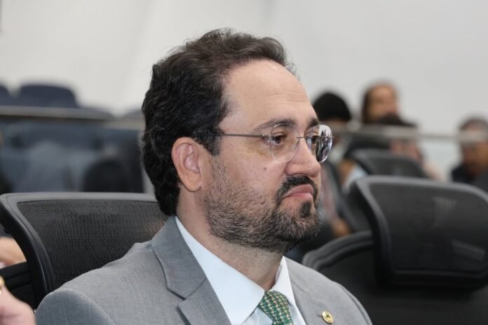 Man in a gray suit and green tie, wearing glasses, listening in a conference room.