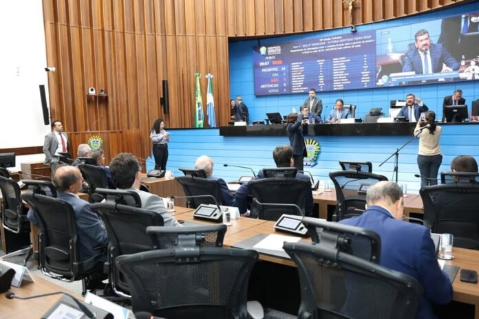 Legislative chamber with officials at curved desks, blue stage, and large screens behind; a speaker addresses the assembly while photographers film.