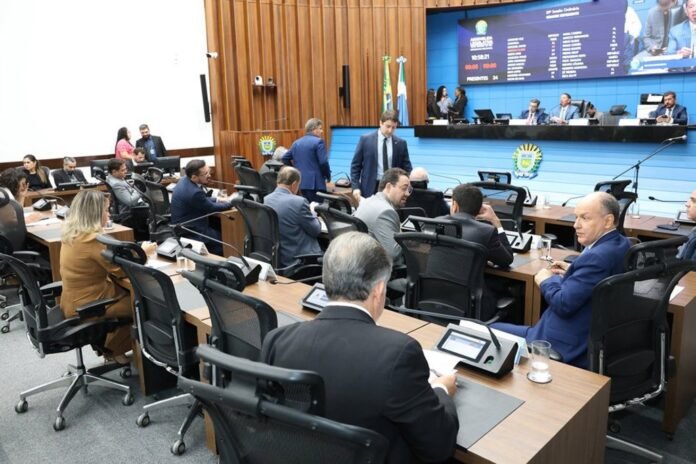 Officials seated at curved desks in a modern government chamber with a large digital scoreboard behind them