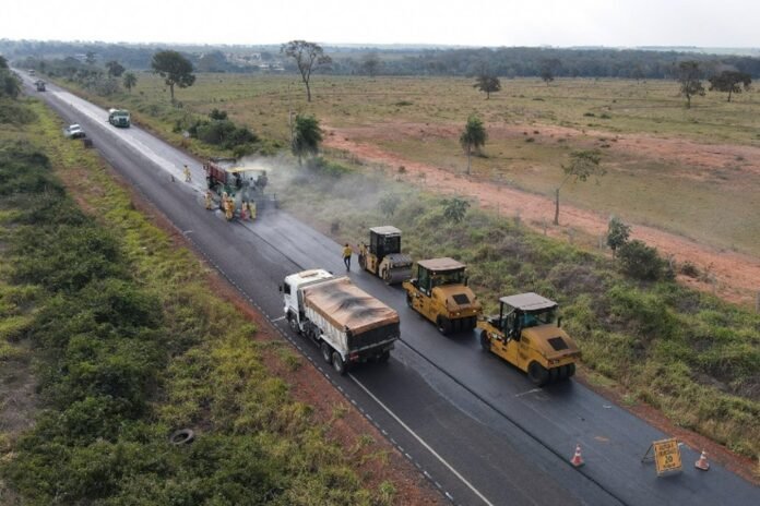 Road construction on a rural highway: workers spread asphalt while heavy rollers compact the surface; a dump truck sits nearby.