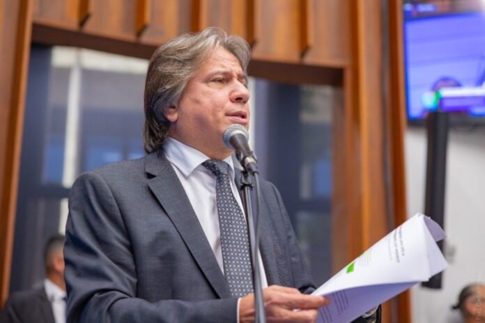 Man in a gray suit and tie speaks into a microphone while holding papers