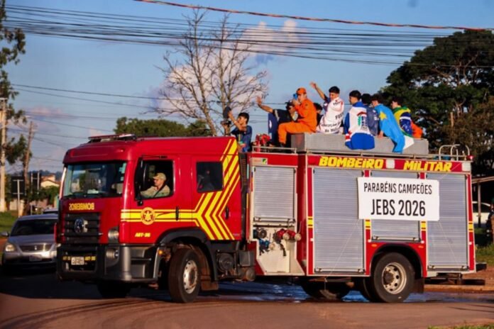 Red fire truck with people on top celebrating in a street parade; banner reads 'Parabéns Campeões JEBS 2026'.