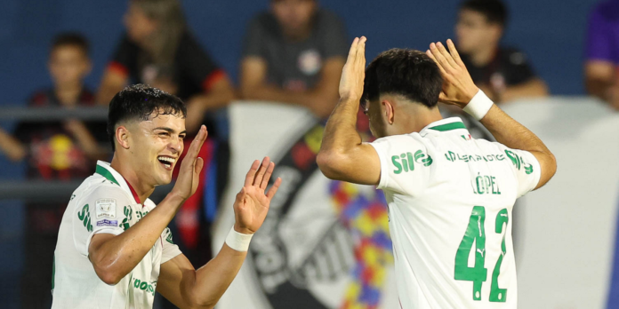 Two soccer players in white and green jerseys celebrate with a high-five on the pitch, smiling after a play, with spectators in the blurred background.