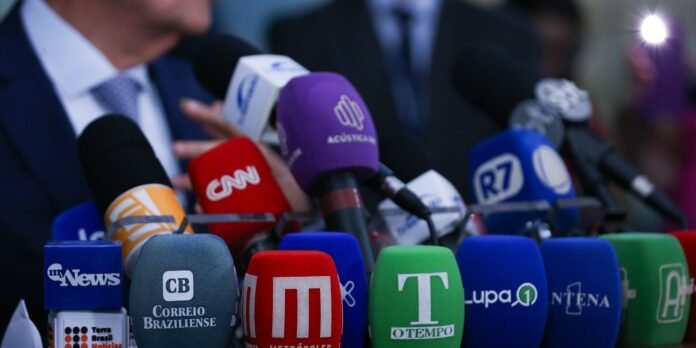 Close-up of a cluster of colorful microphones with news logos at a press conference, in front of a suited speaker.