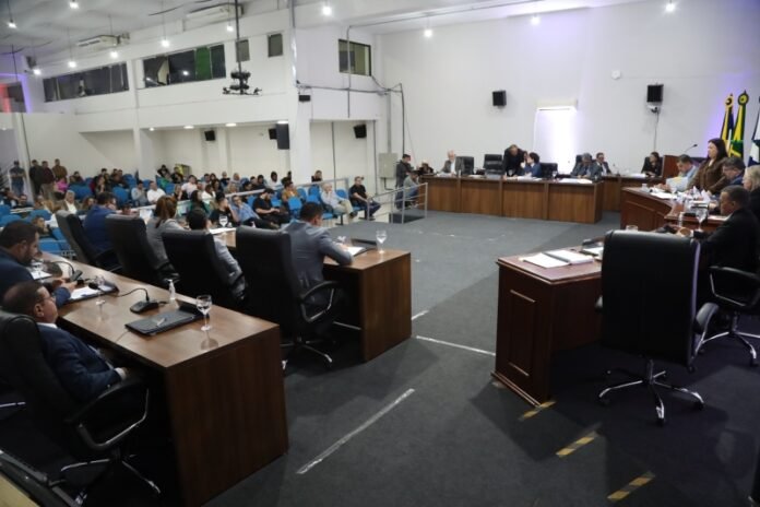 Public council or town hall meeting with a panel at a long desk facing an audience in a large room with microphones and laptops on the desks.