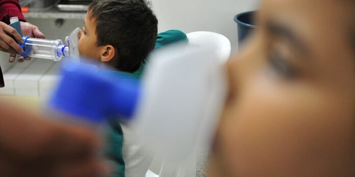 Young boy in a clinic sits in a chair while a caregiver places a clear inhalation mask over his nose and mouth for treatment.