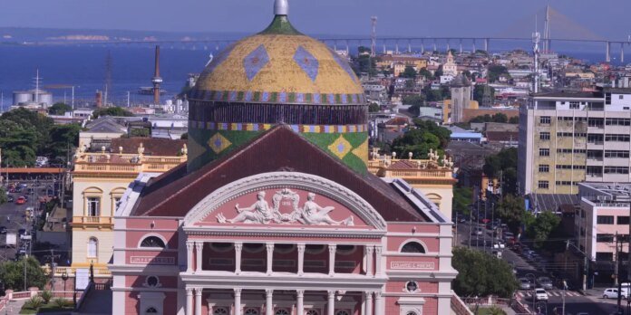 Colorful domed historic building with a patterned tiled dome overlooking a tan-pink neoclassical facade and a busy city street, with a blue bay and long bridge in the background