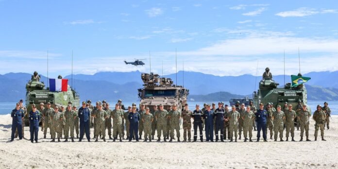 Military personnel in uniform form a long line on a sandy beach with armored vehicles and flags, mountains in the distance with a helicopter overhead.