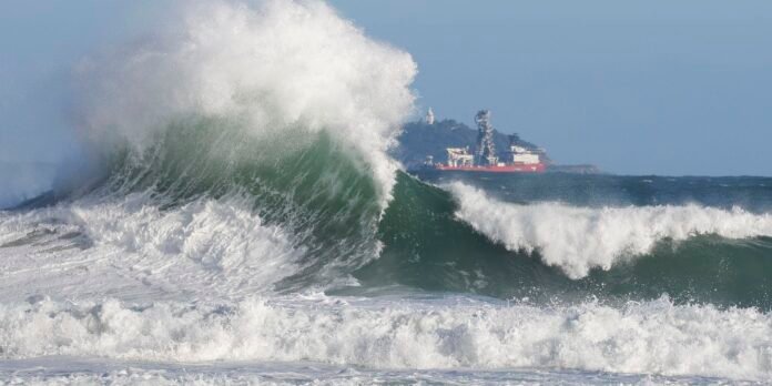 A large curling wave crashes toward the shore with white spray, a red ship visible on the horizon behind the sea spray.