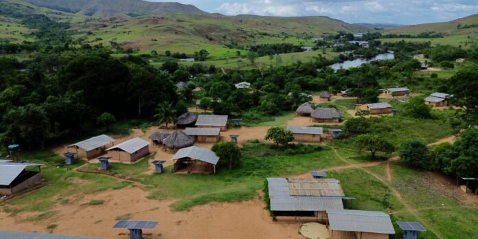 A rural village with thatched huts, metal-roofed shelters, and solar panels in a green valley with hills in the distance.