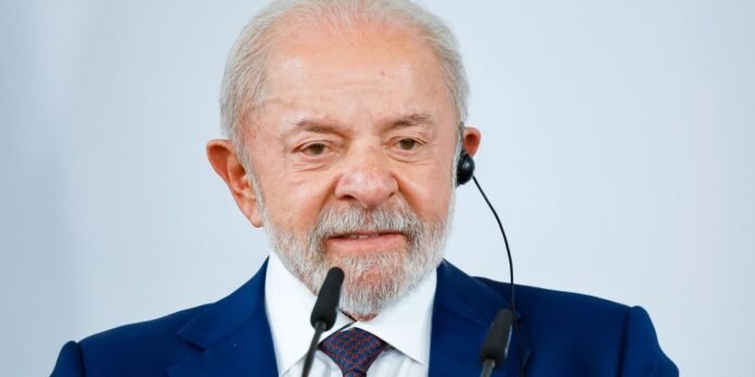 Older man with gray beard in a navy suit speaks into two microphones at a conference podium.