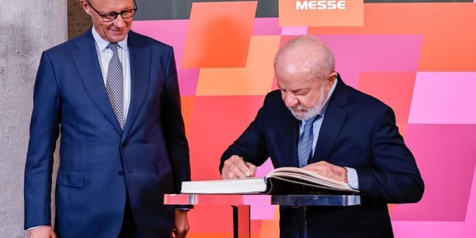 Two men in dark suits at a signing ceremony; one signs a large guest book on a chrome lectern in front of a pink-orange geometric backdrop.
