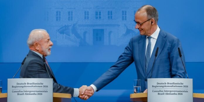 Two suited men shake hands at a blue backdrop during a German-Brazilian government consultations press conference, microphones at the podiums.