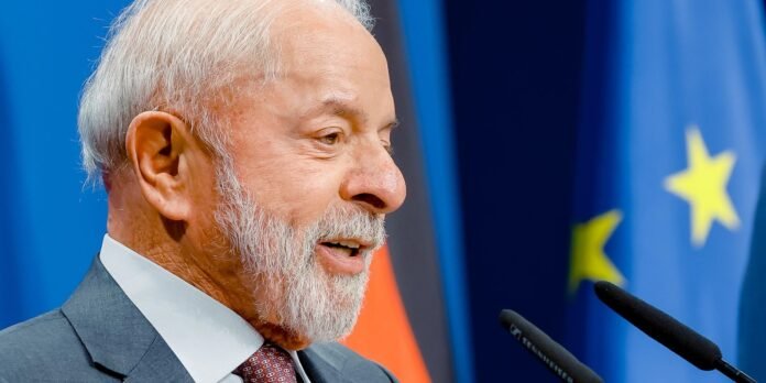 Side profile of an older man with white hair and beard speaking at a podium, EU flag in the background.