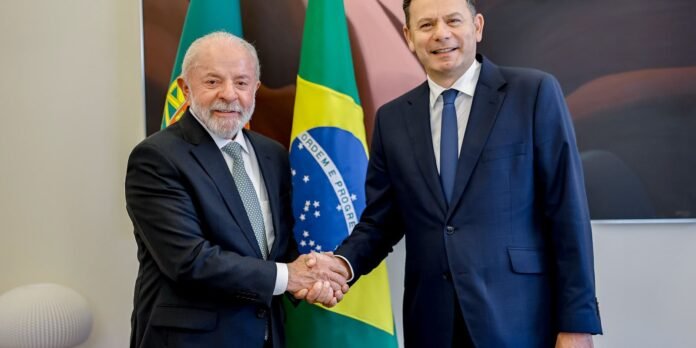 Two officials in suits shake hands in a formal setting, with the Brazilian flag visible behind them.