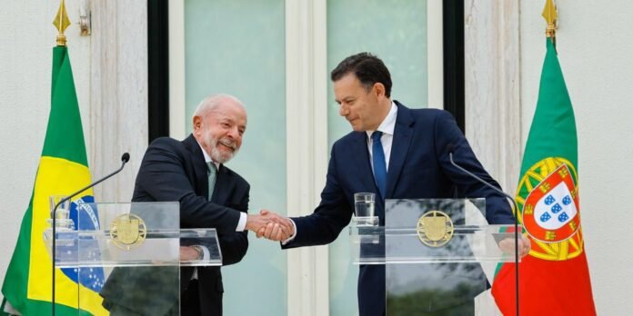 Two men in suits shake hands at podiums during a formal ceremony, with Brazil and Portugal flags in the background.