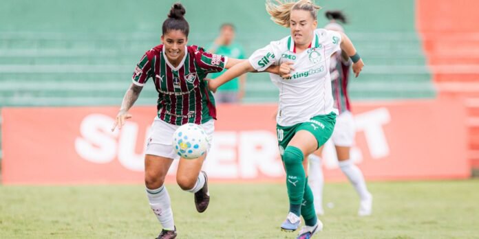 Two female soccer players chase a ball during a match; one wears a red‑green striped kit and the other a white/green kit, with a blurred stadium in the background.