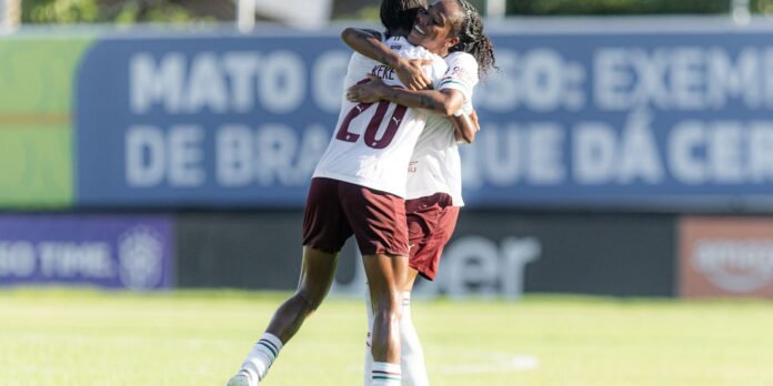 Two soccer players in white and maroon embrace in a celebratory hug on a sunny field, background banners blurred.