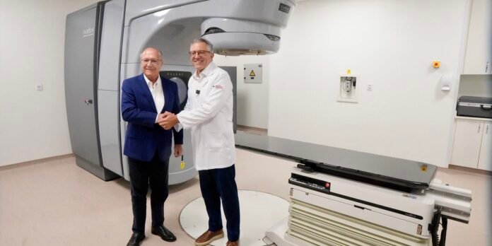 Two men shake hands beside a Varian linear accelerator in a radiotherapy treatment room.
