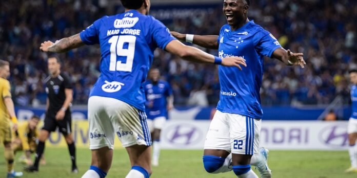 Two blue-jersey soccer players celebrate on a grass field as fans cheer in the stands behind them, arms outstretched.