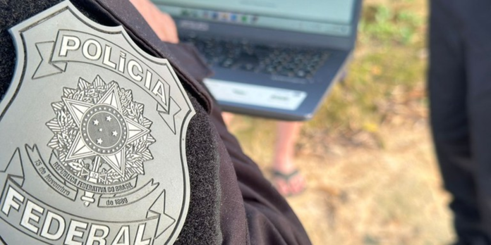 Close-up of a Policia Federal badge pinned to clothing; laptop and outdoor scene blurred in the background.