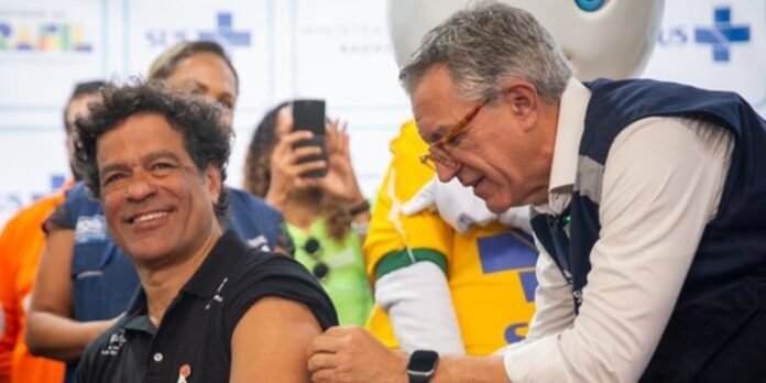 Smiling man in a black polo shirt receives a vaccination from a healthcare worker at a public health event, with onlookers in the background.