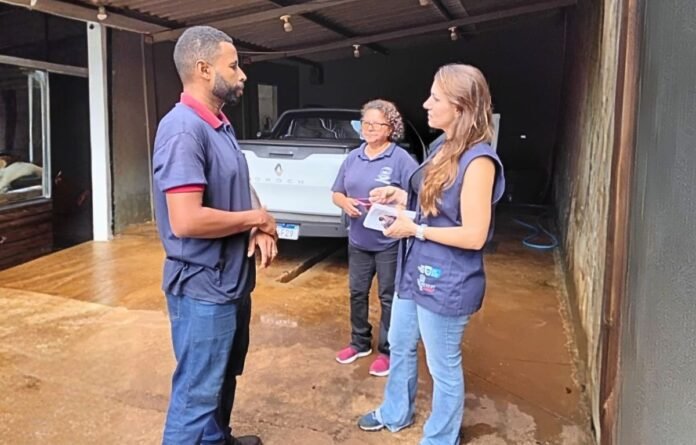 Three people in a car workshop conversing near a light-blue pickup truck inside a garage.