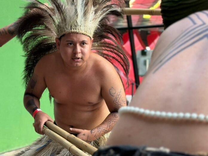 Shirtless man wearing a large feather headdress paddles with two bamboo oars, tattoos on his arm, against a bright green background.