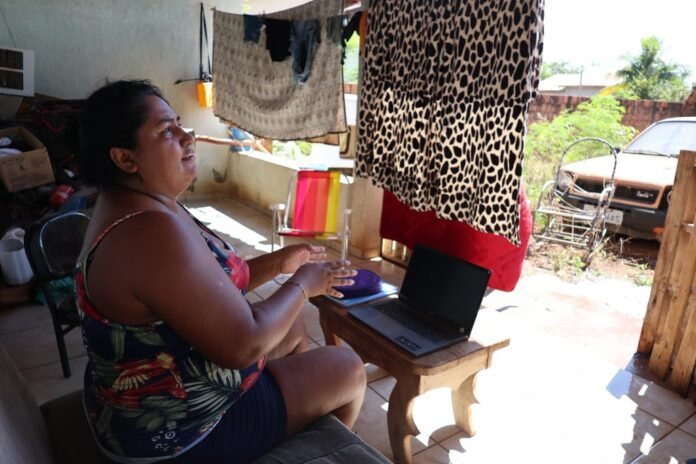 Woman sitting on a porch using a laptop on a small wooden table, with laundry hanging nearby and a car in the yard behind her.
