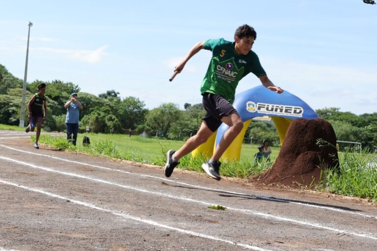 Male runner in a green jersey mid-air over a track line during an outdoor race, with a grassy field and a blue inflatable finish zone in the background on a sunny day