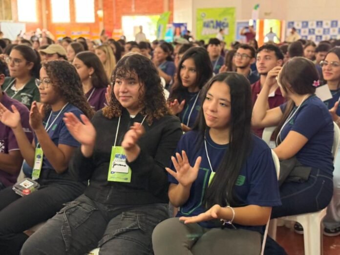 Interior hall with a large audience of young adults seated in rows, many wearing lanyards, clapping during an event.