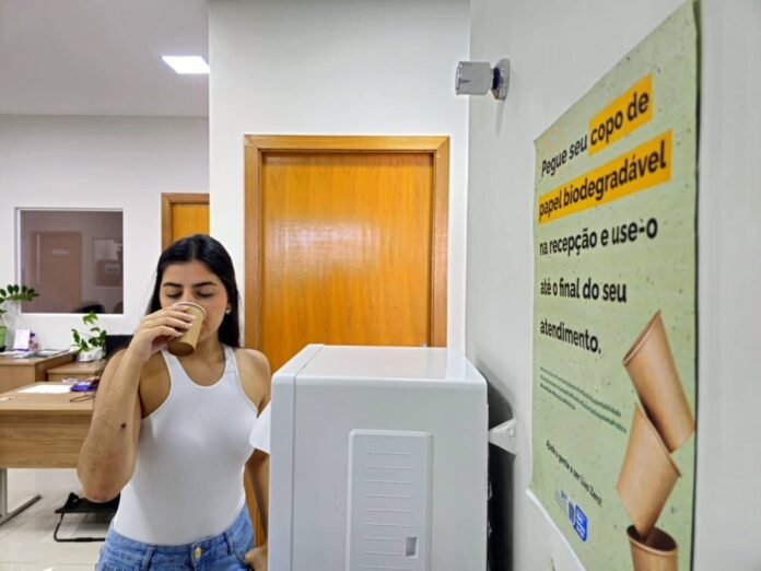 Woman in a white tank top drinks from a paper cup beside a water dispenser in an office systems area.