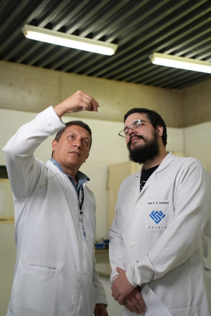 Two scientists in white lab coats examine a small sample held up between them in a laboratory setting.