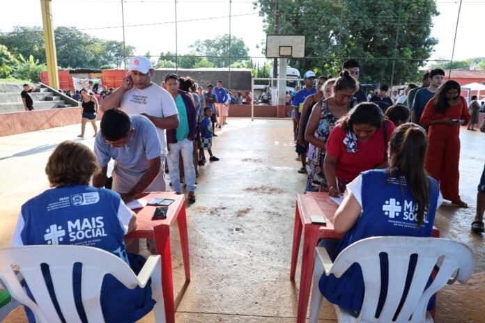 Two volunteers in blue 'Mais Social' vests sit at red tables helping people in a queue at an outdoor community aid event.