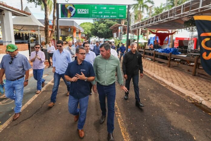 Group of men in smart-casual clothes walking down a street fair, with booths and banners overhead at an expo event.