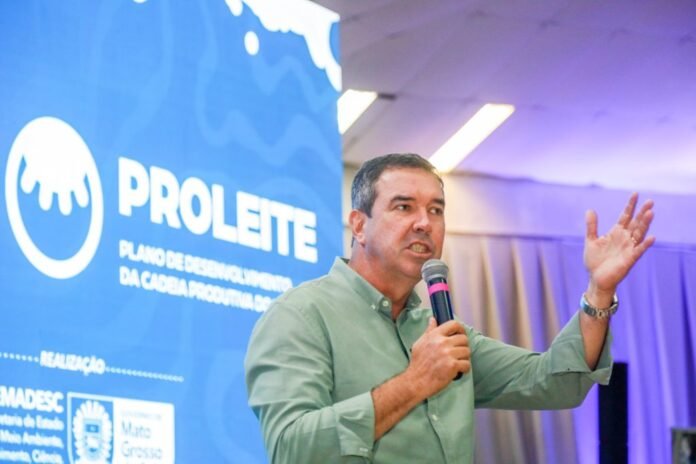 Man in a green shirt speaks into a microphone while gesturing with his left hand on a stage with a blue Proleite banner behind him.
