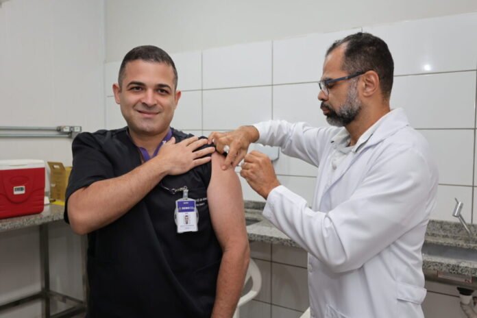 Man in scrubs receives a vaccination from a doctor in a clinical room with tiled walls.