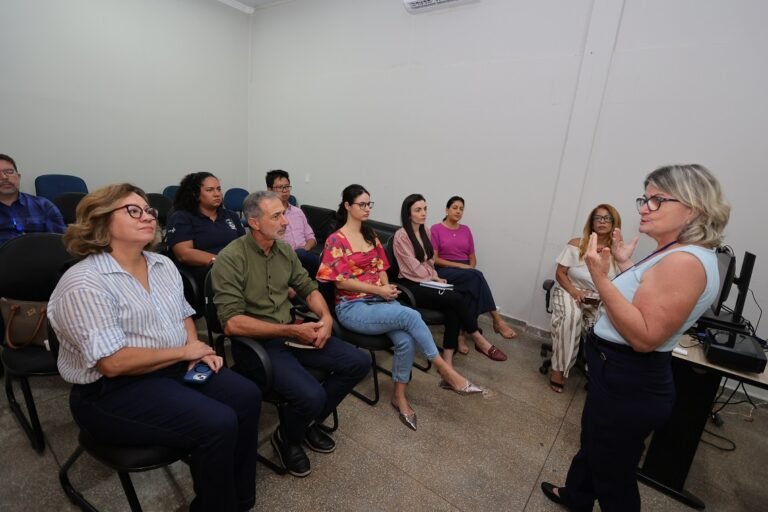 Female presenter in a light blue top speaks to a diverse group seated in a small conference/training room.