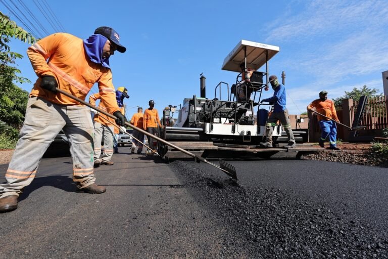 Road construction crew paving fresh asphalt with a paving machine under a clear blue sky is visible creating a new street surface.